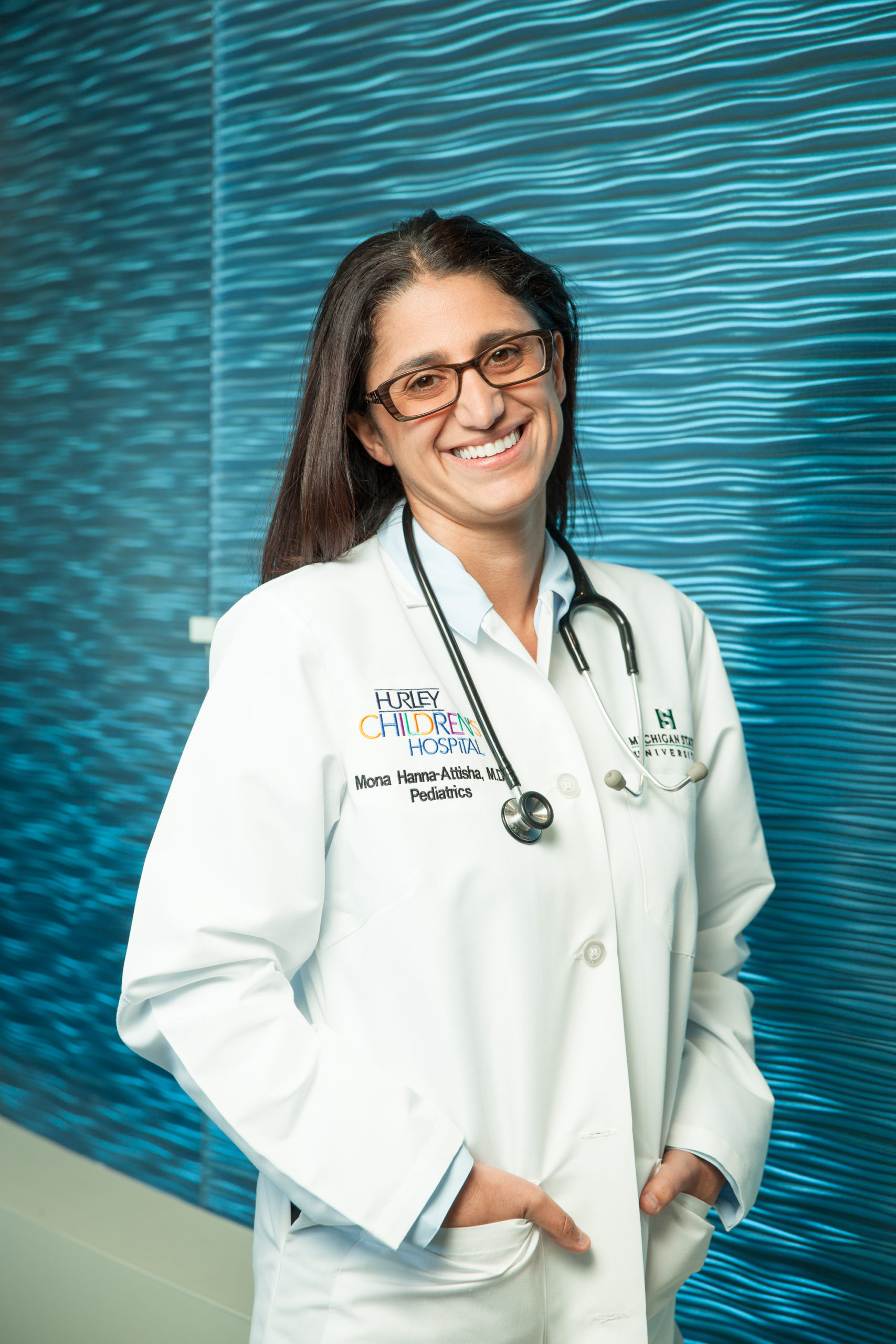 Dr. Mona Hanna-Attisha standing and smiling in front of a blue background